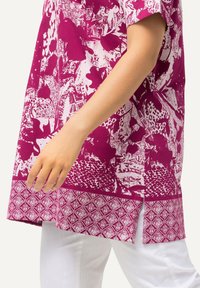 Fuchsia Tunika mit abstrakten weißen Mustern, kurzen Ärmeln und einem gemusterten Saum. Kombiniert mit weißen Hosen, die einen lässigen Schnitt zeigen.