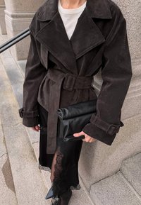 Osoba nosi tmavohnedý semišový kabát so zámkom, pod ktorým má biele tričko a čiernu sukňu s krajkou, drží čiernu koženú kabelku.