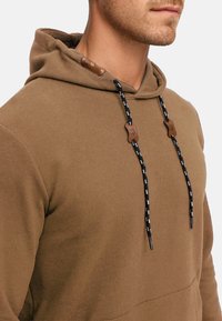 Brun ribbet hoodie med snøre i hætten. Har sort og hvid mønstrede snore med læderdetaljer. Blød tekstur og afslappet design.