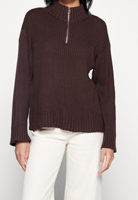 Brauner gerippter Pullover mit hohem Kragen und Reißverschluss bis zur Hälfte, mit langen Ärmeln und entspannter Passform; getragen mit cremefarbenen Hosen.