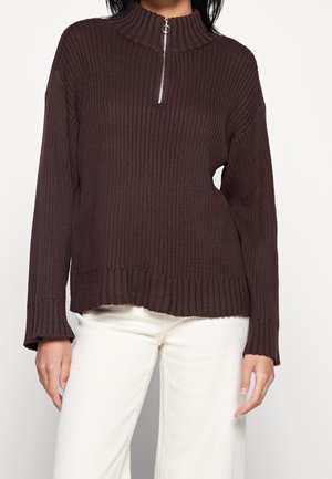 Brun ribbet sweater med høj krave og halvlukning, med lange ærmer og en afslappet pasform; båret med cremede bukser.