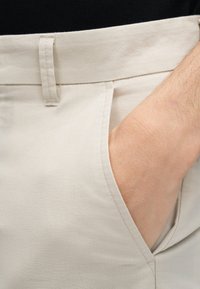 Χέρι που εισέρχεται μερικώς στην μπροστινή τσέπη ανοιχτό μπεζ παντελονιού με ορατό θηλάκι ζώνης και ραφή.
