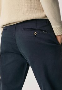 Mörkblå byxor gjorda av en bomullsblandning med en mjuk textur, med två bakfickor och en knappdetalj, stylade med en ljusbeige tröja.