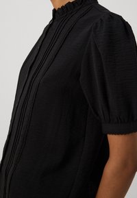 Blusa negra con un escote con volantes, mangas cortas abullonadas, tejido texturizado y detalles de pliegues en la parte delantera.