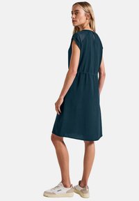Dunkelblaugrünes Kleid mit kurzen Ärmeln und taillierter Taille, mit einem Schlüsselhalo-Ausschnitt hinten. Kombiniert mit weißen und braunen Sneakers.
