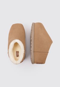 Zoccoli in suede beige con fodera in shearling, punta arrotondata, suola piatta e dettaglio del logo stampato sul tallone.
