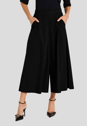 Schwarze weitgeschnittene Hosen mit Falten, hohem Taillenbund und seitlichen Taschen, kombiniert mit schwarzen Absatzschuhen und einem figurbetonten Oberteil. Glatte Textur.