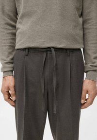 Sivkasti pleten pulover z rebrastimi rokavi, oblečen preko temnosivih hlač z vrvico v pasu in gladko teksturo blaga.