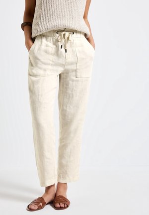 Pantalones beige con cordón y bolsillos delanteros, usados con un top de punto sin mangas y sandalias planas marrones. Manos en los bolsillos, de pie sobre fondo blanco.