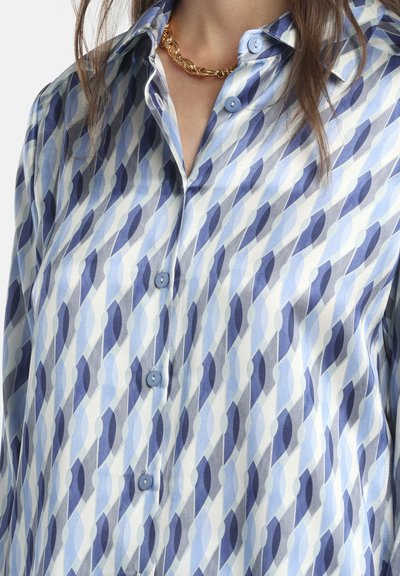 Camicia di seta con motivo geometrico in tonalità di azzurro chiaro e scuro. Colletto con bottoni e due bottoni azzurri visibili.