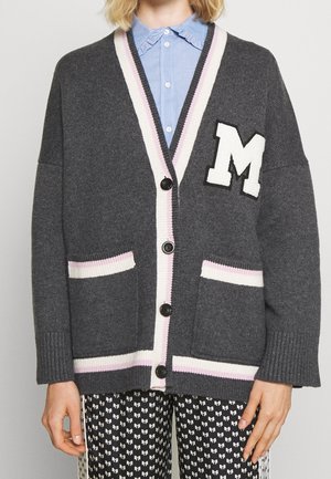 Cardigan gris boutonné avec bordure rayée blanche et rose, grand écusson blanc en forme de lettre M, porté sur une chemise bleue à col et un pantalon noir à motifs.