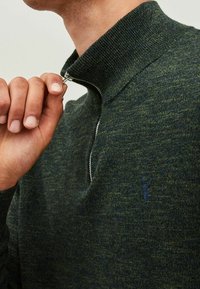 Tmavě zelený pletený pulovr s vysokým límcem a stříbrným zipem. Má jemnou strukturu a malou logo na hrudi.