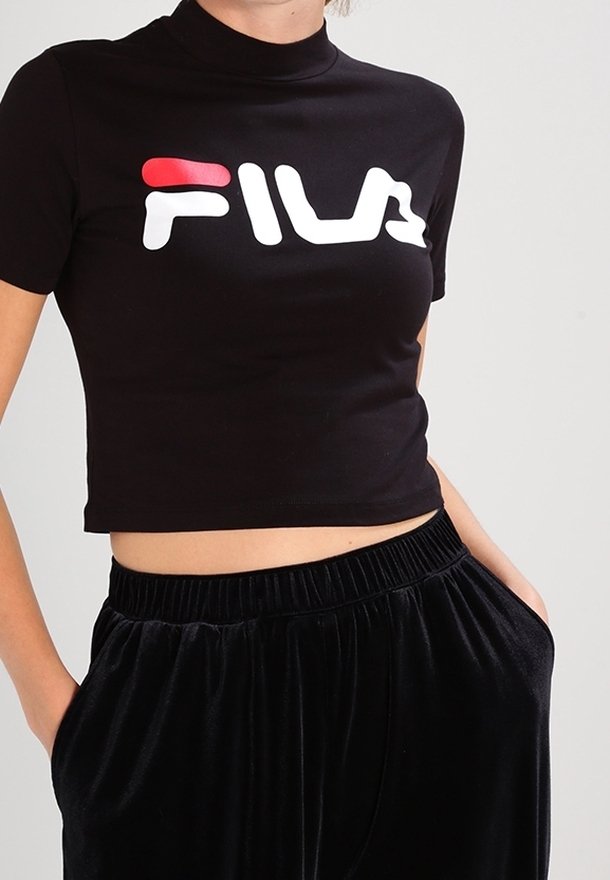 Personne portant un t-shirt court noir avec un grand logo FILA blanc et rouge et un pantalon noir ample, les mains dans les poches.