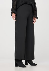 Pantaloni neri a gamba larga realizzati in un tessuto morbido, con vita alta, abbinati a stivaletti neri con tacco. Blusa nera testurizzata con polsini volants.