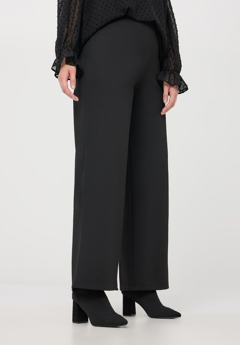 Pantaloni neri a gamba larga realizzati in un tessuto morbido, con vita alta, abbinati a stivaletti neri con tacco. Blusa nera testurizzata con polsini volants.