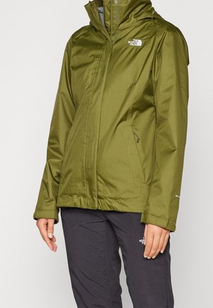 Person iført en olivengrøn vandtæt jakke og sorte bukser med The North Face-logoer synlige på begge.