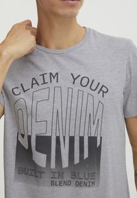 Grå bomulls t-skjorte med stor svart tekst: "CLAIM YOUR DENIM" og "BUILT IN BLUE." Korte ermer, rund hals, slank og glatt tekstur.