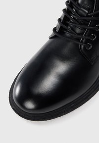 Botín de cuero negro con un acabado suave, puntera redonda y cordones negros en contraste pasados por ojales metálicos. Suela de goma.