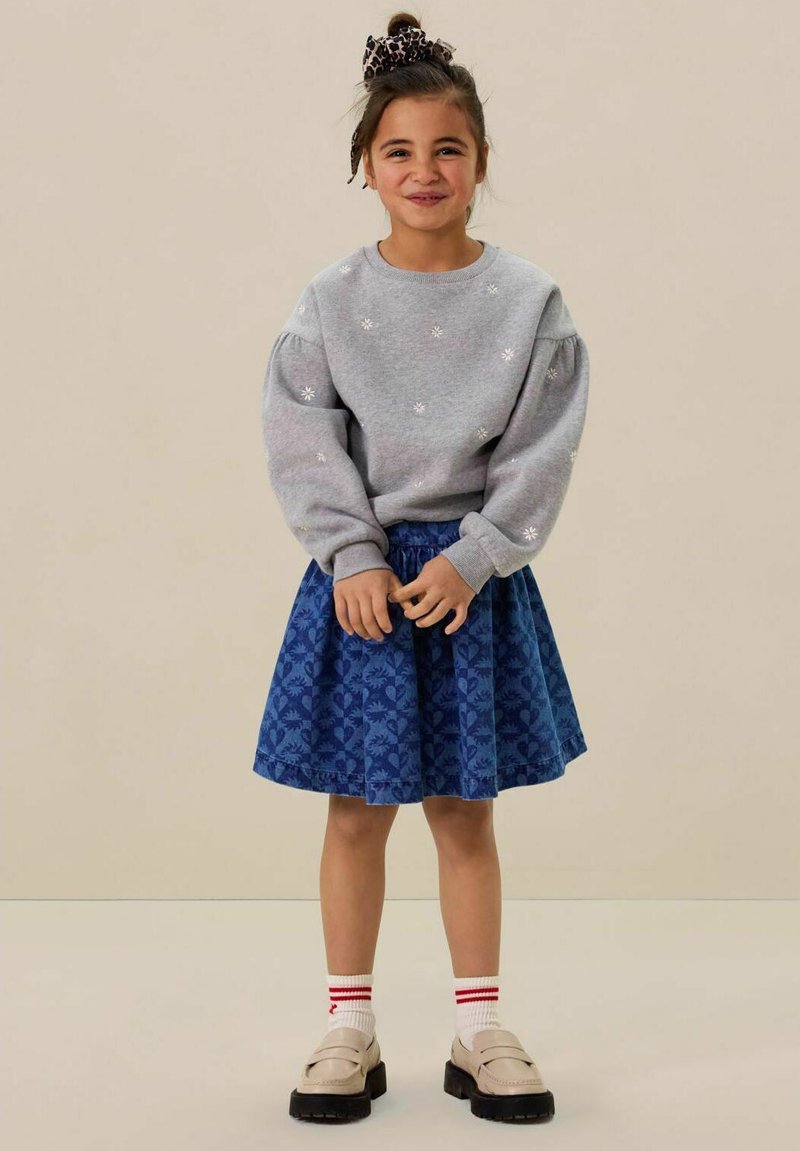 Sweat-shirt gris avec broderie florale, associé à une jupe bleue présentant une texture à motifs. Complété par des chaussettes blanches et des chaussures noires.