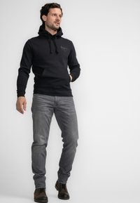Sudadera negra con bolsillo tipo canguro, capucha ajustable y detalle de logo. Combinada con jeans grises y botas marrones con cordones. Fondo sencillo.