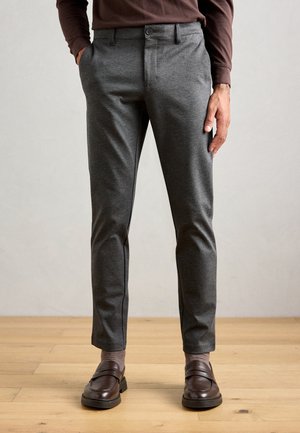 Man med slim-fit grå byxor, bruna strumpor, mörkbruna lädersandaler och en brun skjorta med långa ärmar står på trägolv.