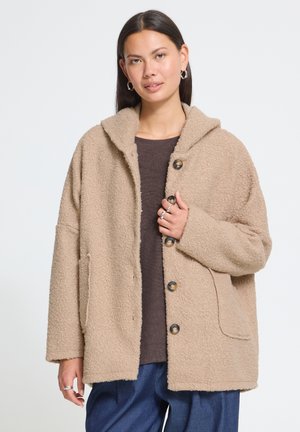 Femme aux cheveux foncés portant des boucles d'oreilles argentées en forme d'anneau, un manteau teddy beige boutonné sur un haut marron et un pantalon bleu sur un fond uni.