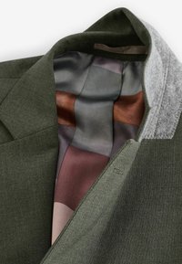 Blazer vert olive avec un tissu extérieur lisse et texturé. L'intérieur présente une doublure en soie à motifs dans des tons sourds de gris, marron et bordeaux.