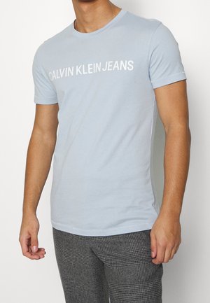 Jasnoniebieska bawełniana koszulka z krótkimi rękawami i okrągłym dekoltem, z napisem "CALVIN KLEIN JEANS" w kolorze białym na piersi.