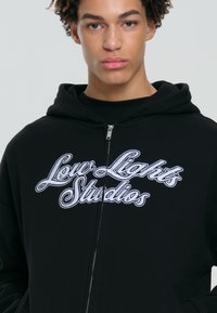 Giovane con capelli ricci che indossa una felpa nera con zip e testo bianco "Low Lights Studios" sul petto.