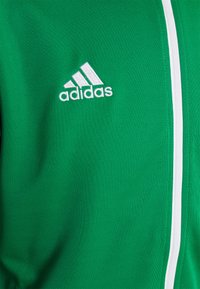 Grønn Adidas-jakke med hvit glidelås og logo, laget av teksturert stoff, med distinkt søm og rene linjer.