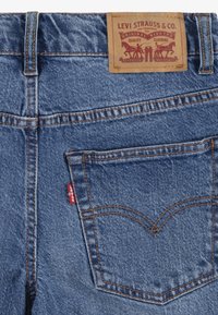 Blå denimjeans med en brun läderlapp som har Levi Strauss-logotypen, kopparfärgade sömmar och en sidofickan som framhävs av en röd etikett.