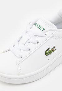 Sneaker bianco con tomaia in sintetico testurizzato, punta rotonda e lacci piatti. Presenta il logo Lacoste verde e un ricamo di coccodrillo sul lato.