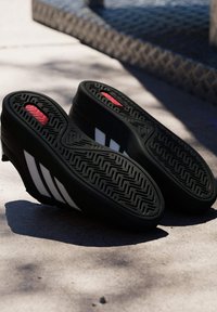 Paire de crampons noirs avec des bandes blanches et des détails rouges, semelles vers le haut sur un sol en béton ensoleillé avec des ombres.