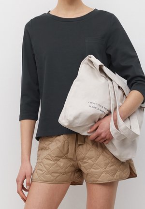 Osoba ubrana w czarną bluzkę z długim rękawem i beżowe pikowane szorty, trzymająca złożoną beżową torbę na ramię z napisem.