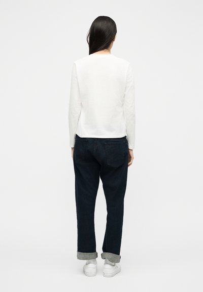 Γυναίκα με μακριά σκούρα μαλλιά, φορώντας λευκό πουκάμισο με μακριά μανίκια, σκούρα τζιν με ρεβέρ και λευκά αθλητικά παπούτσια, όρθια με πλάτη σε απλό φόντο.