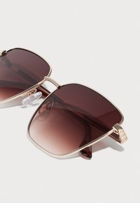 Quadratisch geformte Sonnenbrille mit einem goldenen Metallrahmen. Dunkelrot verlaufende Gläser mit transparenten Nasenpolstern und dekorativen Seitenakzenten.