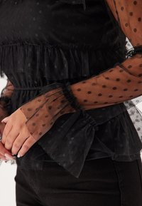 Svart polka prickig meshblus med volangdetaljer, långa genomskinliga ärmar och samlad midja. Texturerad tyg ger djup och kontrast i mönstret.