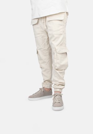 Pantalons cargo beige avec des poignets élastiques et plusieurs poches, associés à des baskets en daim gris clair avec des semelles et des lacets blancs.