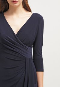 Nainen, jolla on päällään tummansininen kietaisumekko, jossa on kolmen neljänneksen pitkät hihat ja kootut kankaat vyötäröllä ja hameessa.