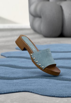 Egyetlen fa szandál farmerral pánttal és fém szegecsekkel, egy texturált kék szőnyegen, háttérben elmosódott karosszékkel.