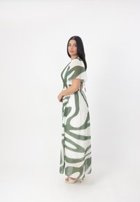 Robe maxi en tissu léger, présentant une base blanche avec des motifs abstraits verts. Les manches courtes et une taille ajustée soulignent la silhouette.