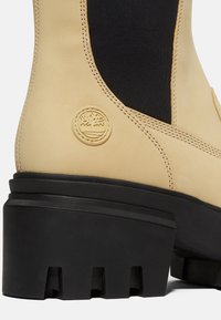 Botte beige avec semelle épaisse noire à crampons, panneau latéral élastique noir, coutures détaillées et logo rond en relief d'arbre sur la zone du talon.