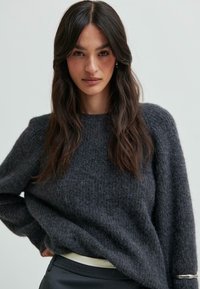 Mladá žena s dlouhými tmavými vlasy ve svetru z textured pleteniny v antracitově šedé barvě a černých kalhotách, pózující s rukou na boku před jednobarevným pozadím.