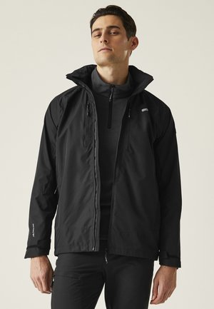 Mężczyzna ubrany w czarną, outdoorową kurtkę z zamkiem na zewnątrz, na czarnej polarowej bluzie z wysokim kołnierzem, na tle jasnego, jednolitego tła.