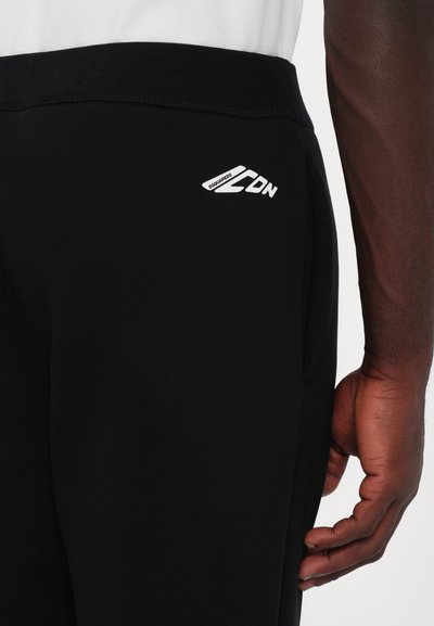 Schwarze Sporthosen mit glatter Textur, ausgestattet mit einem weißen Logo an der Seite und einem enganliegenden Bund. Seitliche Taschen sind sichtbar.