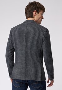 Dunkel gemusterter Blazer aus strukturiertem Stoff, mit Reverskragen, einzelner Rückenbelüftung und knöpfbaren Manschetten. Trägt dazu eine dunkle Jeans.