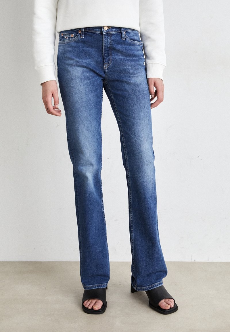 Personne portant un jean bleu droit, un haut blanc à manches longues et des sandales noires à talons ouverts, debout sur un sol clair.