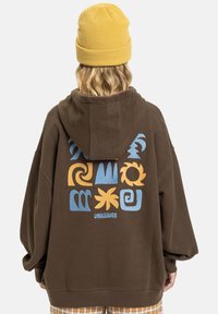 Brauner oversized Hoodie mit einem großen Grafikdruck in blauen und orangefarbenen Mustern auf dem Rücken. Kombiniert mit einer gelben Strickmütze.