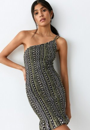 Mujer con un vestido ajustado sin mangas y de un solo hombro con rayas verticales en negro, verde y blanco, de pie contra una pared blanca.