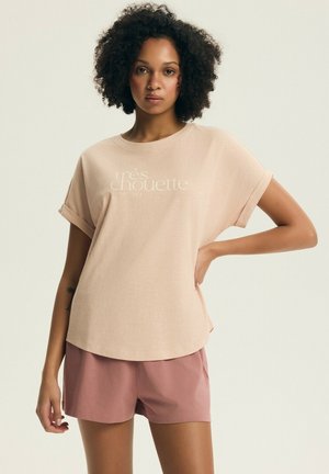 Kobieta z kręconymi włosami w beżowej, obszernej koszulce z napisem i brudnoróżowych szortach, stojąca z jedną ręką na biodrze.